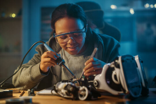 Young Teenage Multiethnic Schoolgirl is Studying Electronics and Soldering Wires and Circuit Boards in Her Science Hobby Robotics Project. Girl is Working on a Robot in Her Room. Education Concept.